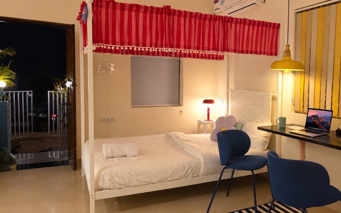 A studio room at night: a white four-poster bed with a red canopy, a desk with blue chairs, and the door open to the landing outside