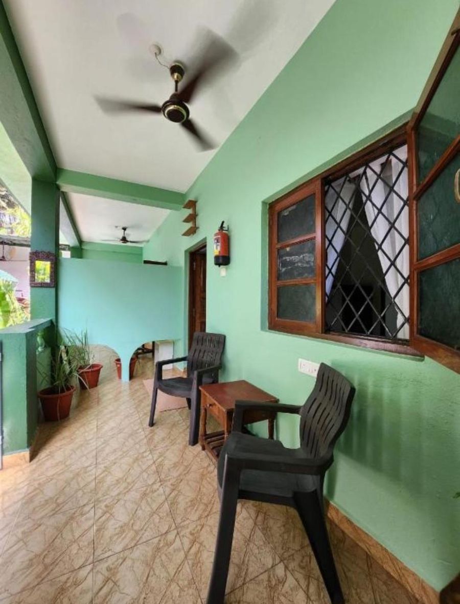 A covered balcony with two chairs and a small wooden table beside a room window, under a ceiling fan