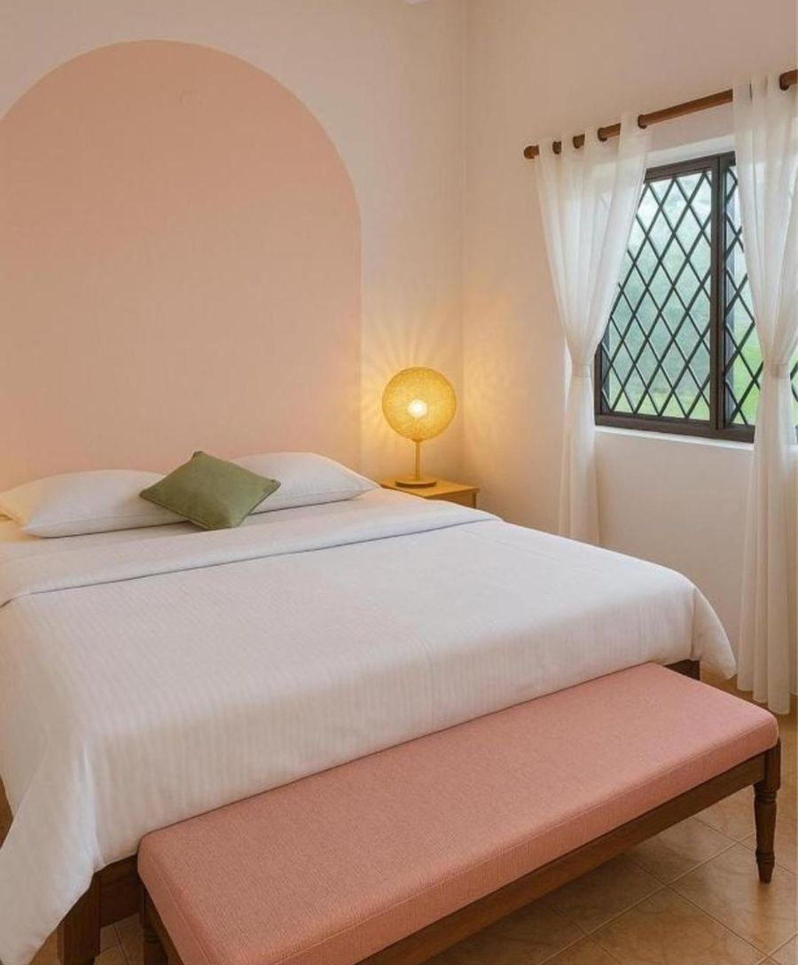 A bedroom with a pink arched headboard wall, a white double bed, a pink bench at the foot and a glowing globe lamp by the window