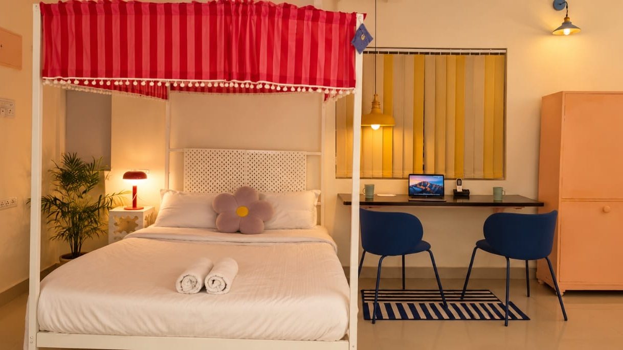 A studio room with a white four-poster bed under a red striped canopy, a wall desk with two blue chairs and a peach wardrobe