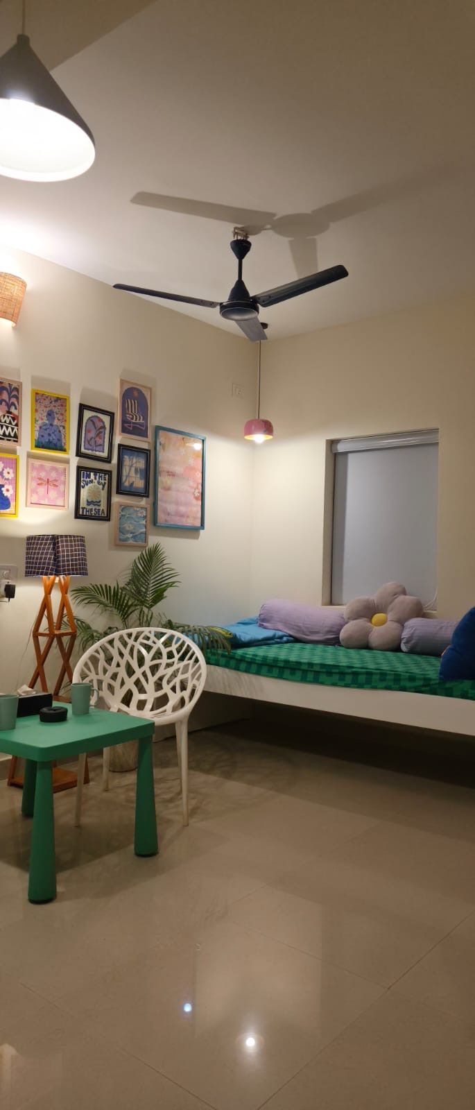 A day-bed with a green mattress and cushions beneath a gallery wall of framed prints, with a green table and a white chair