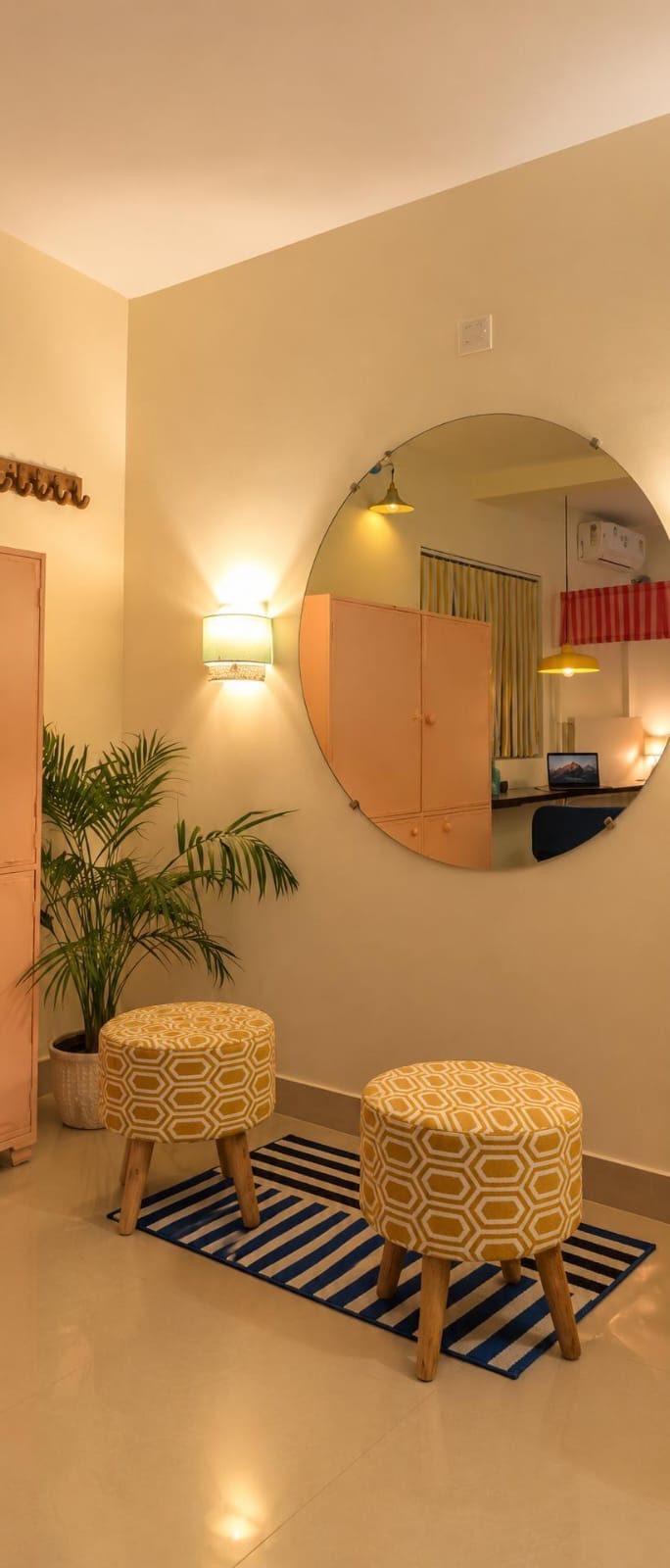 A round wall mirror above two mustard pouffes on a blue striped rug, beside a potted palm and a wall lamp